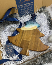 A wooden Christmas tree-shaped cheese board with a beach coloured rolling resin waves, presented in a gift box with shredded paper, silver tinsel, blue ribbons, and a brand card.