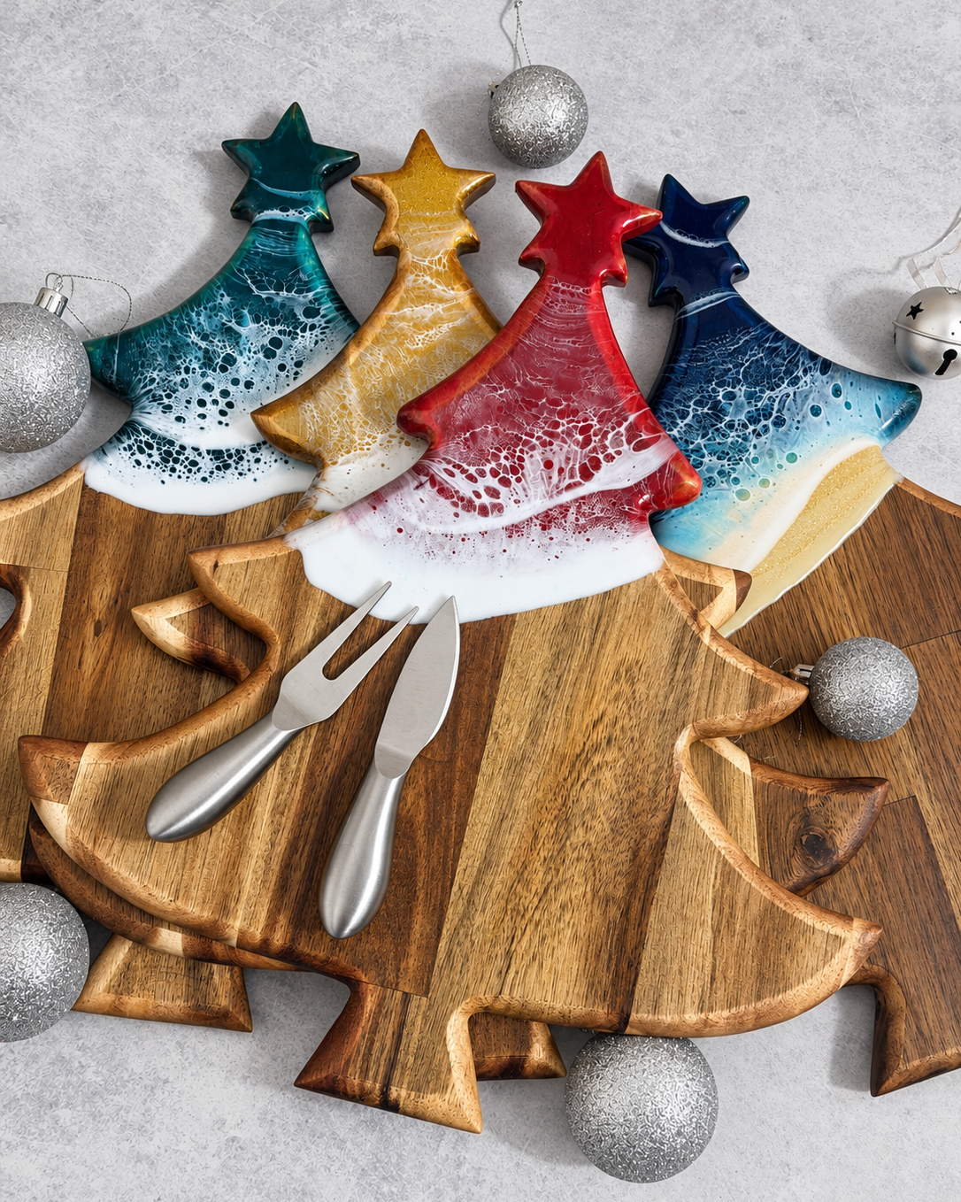 A set of Christmas tree-shaped wooden serving boards in red, blue, green and gold, styled with silver serving utensils and silver glitter ornaments on a gray textured background.