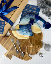 A holiday-themed gift set featuring a wooden Christmas tree-shaped serving board with a blue and cream ceramic star-shaped cheese board on top, accompanied by two silver spoons, a kraft gift box with a blue ribbon, and a product card from Element6.