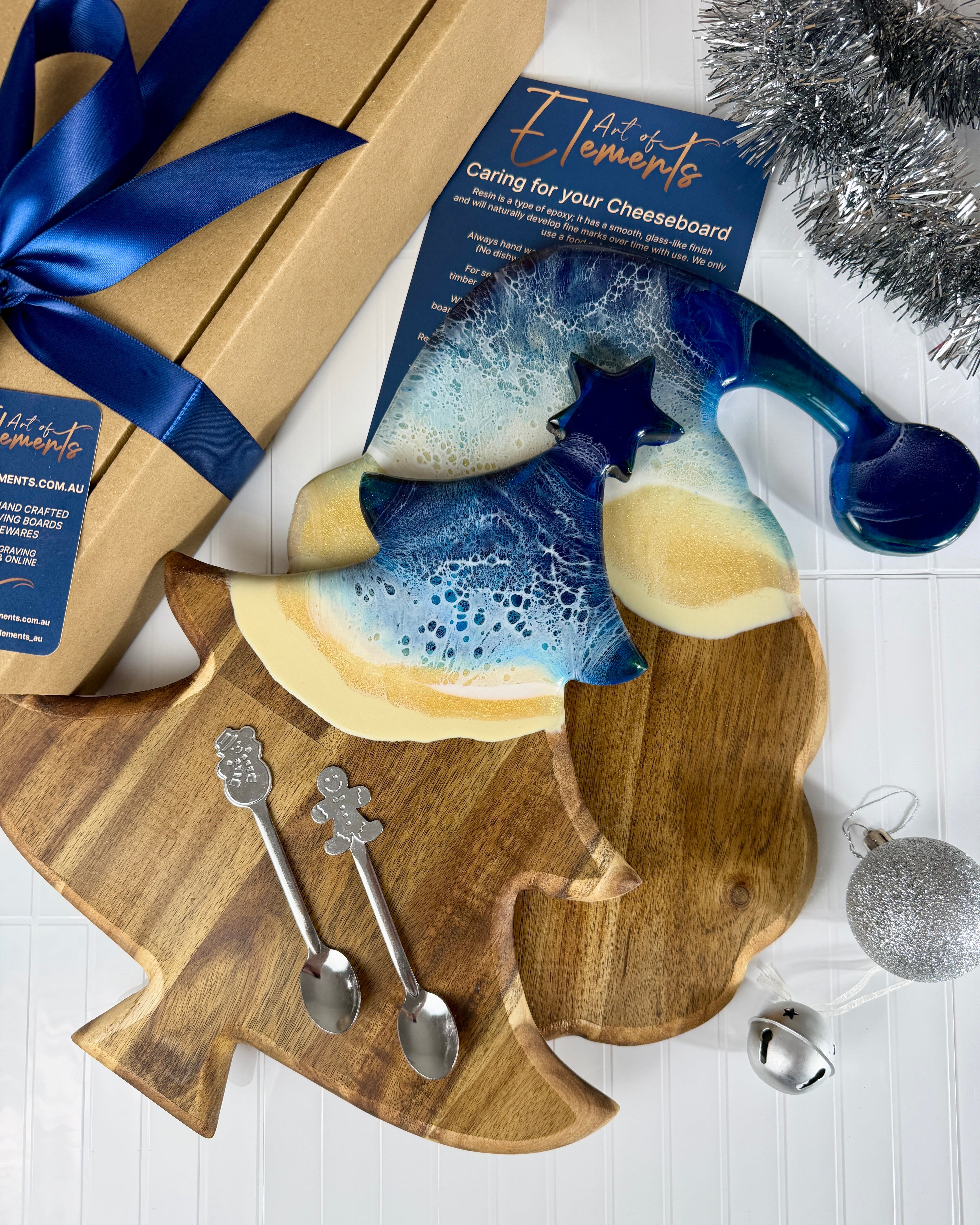 A holiday-themed gift set featuring a wooden Christmas tree-shaped serving board with a blue and cream ceramic star-shaped cheese board on top, accompanied by two silver spoons, a kraft gift box with a blue ribbon, and a product card from Element6.