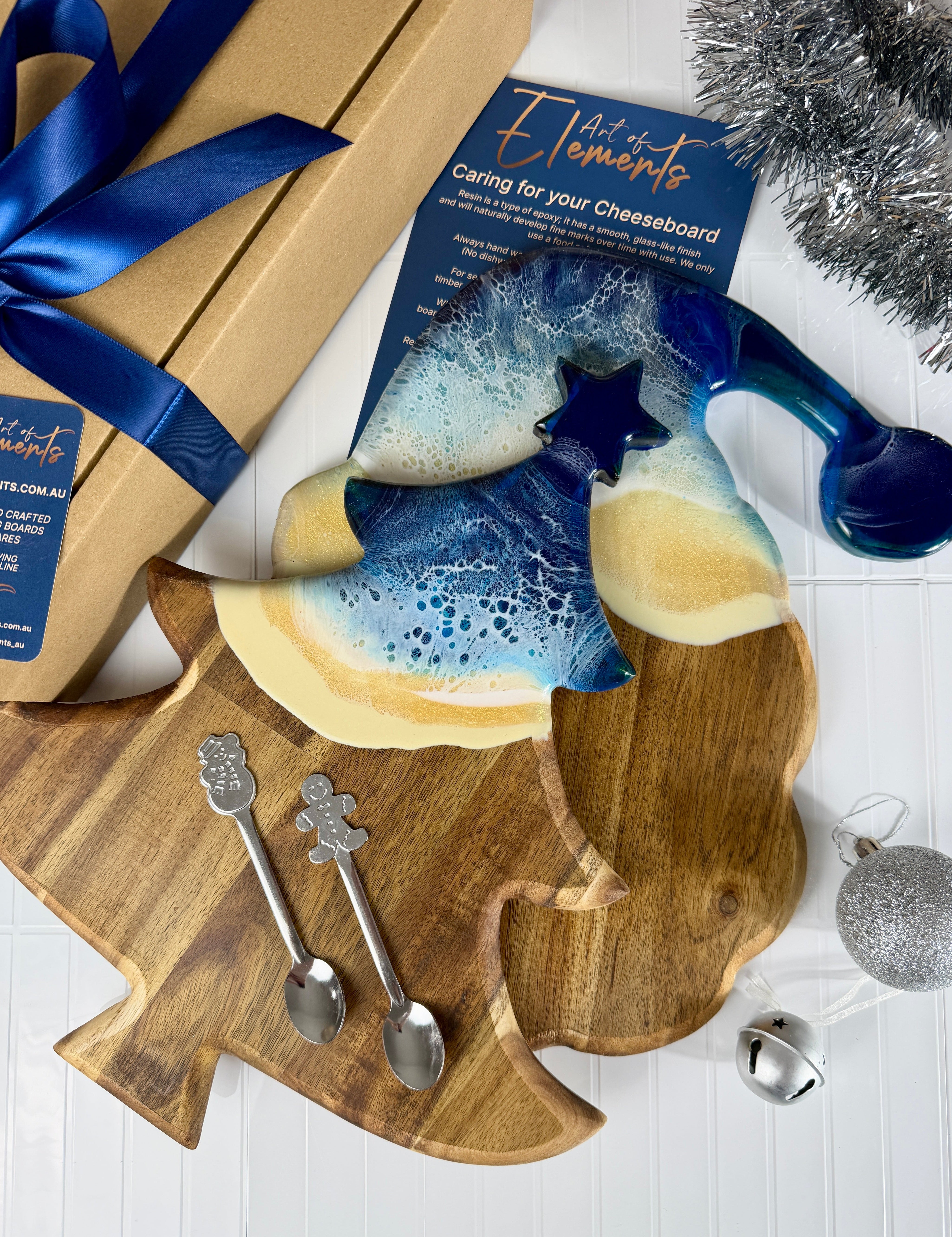 A holiday-themed gift set featuring a wooden Christmas tree-shaped serving board with a blue and cream ceramic star-shaped cheese board on top, accompanied by two silver spoons, a kraft gift box with a blue ribbon, and a product card from Element6.