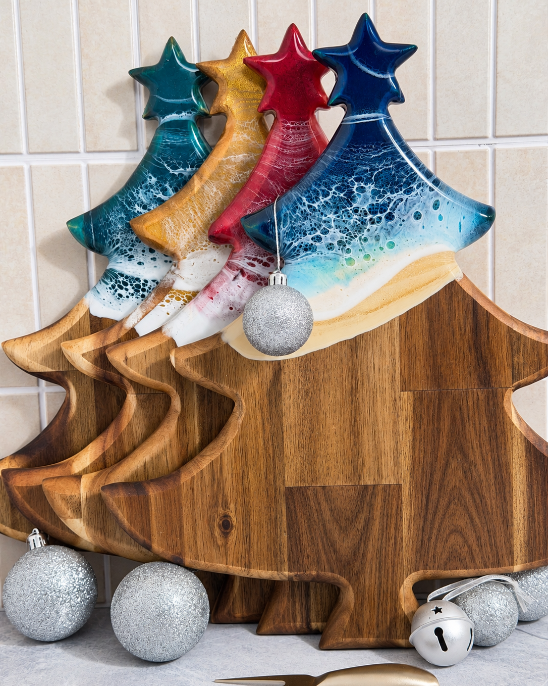 A Christmas-themed wooden serving board shaped like a tree, with rolling resin waves in, green, gold, red, and beach, along with silver glitter balls and a small silver bell, set against a tiled kitchen backsplash.