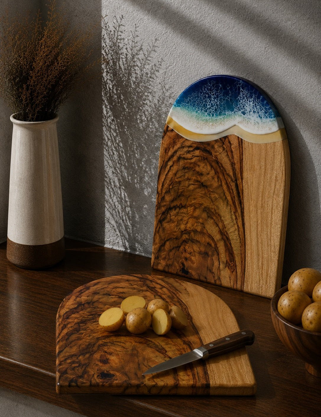 One chopping board laying flat on the kitchen bench with cut up potatoes, and the same shaped arch board with the beach resin waves leaning up against the kitchen wall.