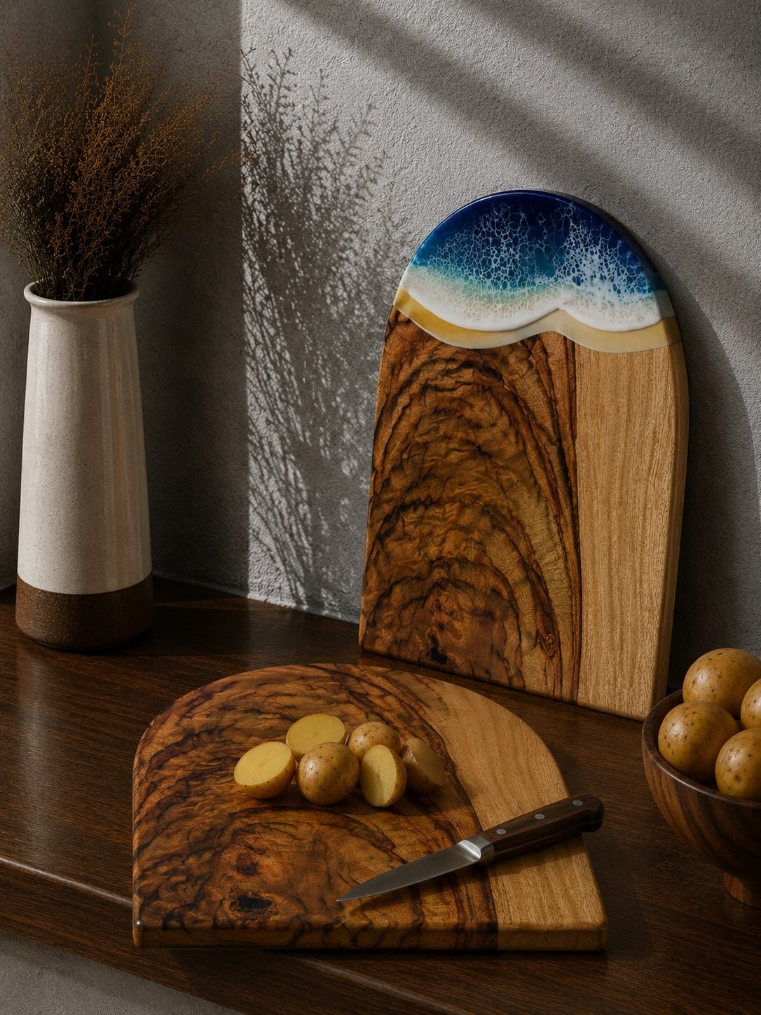 One chopping board laying flat on the kitchen bench with cut up potatoes, and the same shaped arch board with the beach resin waves leaning up against the kitchen wall.