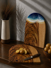 One chopping board laying flat on the kitchen bench with cut up potatoes, and the same shaped arch board with the beach resin waves leaning up against the kitchen wall.