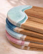 A stack of round wooden serving boards with ocean-wave cutouts, featuring natural camphor laurel wood grain and coloured wave designs in light blue, mint green, pink, beige and beach colourways  