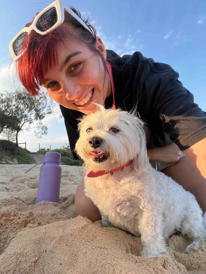 Woman with red hair and white sunglasses kneeling on sand with a small white dog and a purple water bottle