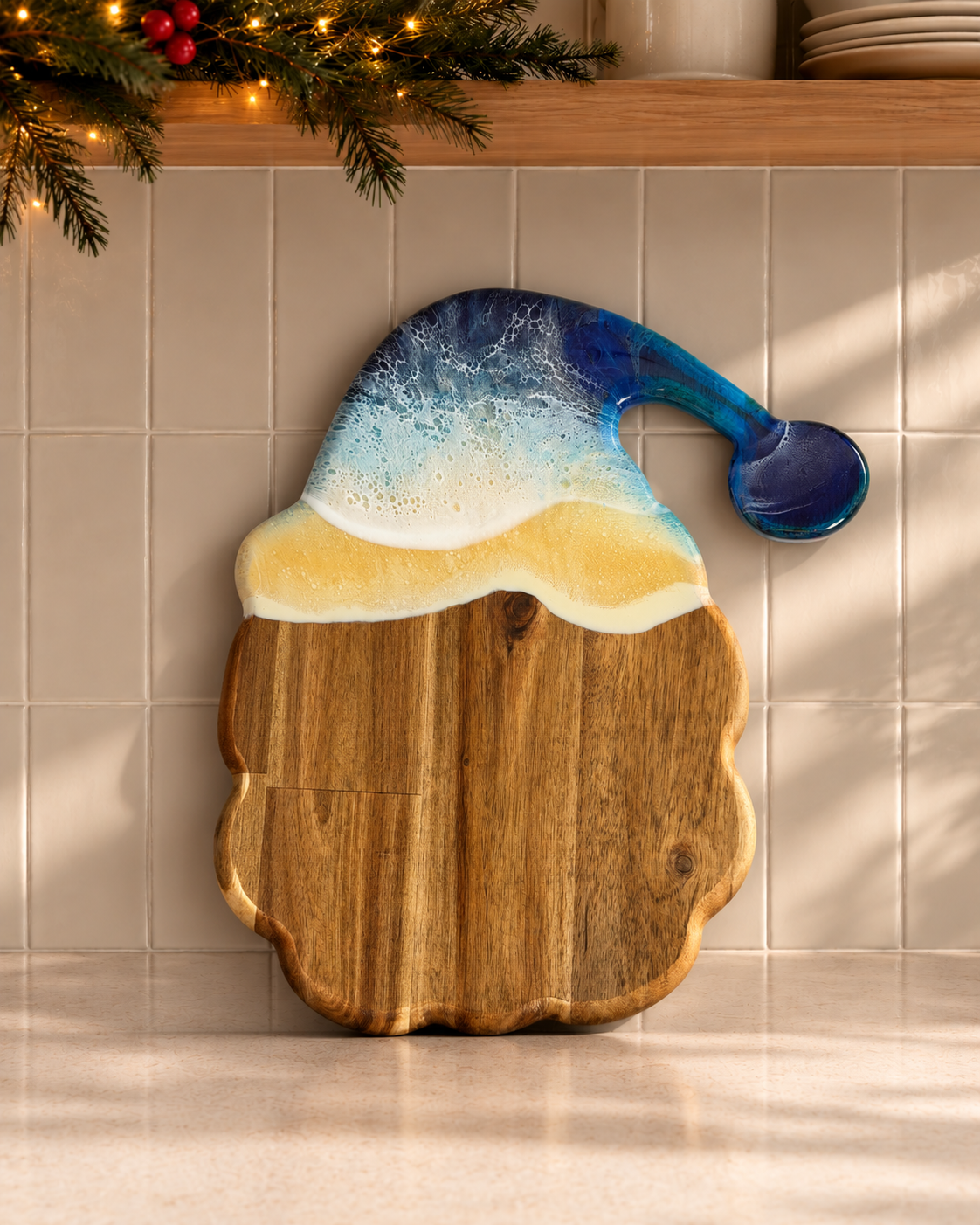 A wooden holiday cutting board shaped like a Santa hat, with a natural wood body and a resin top featuring blue, white, and yellow beach-inspired swirls, displayed against a beige tiled wall with holiday greenery above.