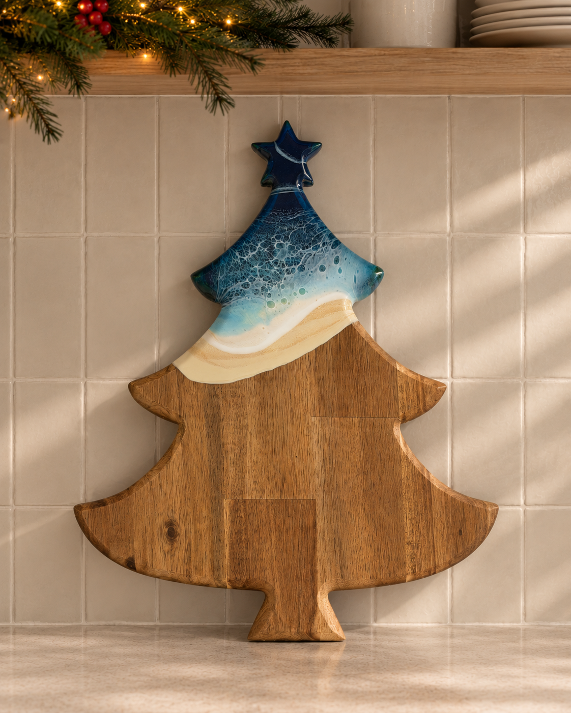 A Christmas tree-shaped wooden cutting board with a painted resin design of a blue ocean wave and sandy beach, leaning against a beige tiled wall.