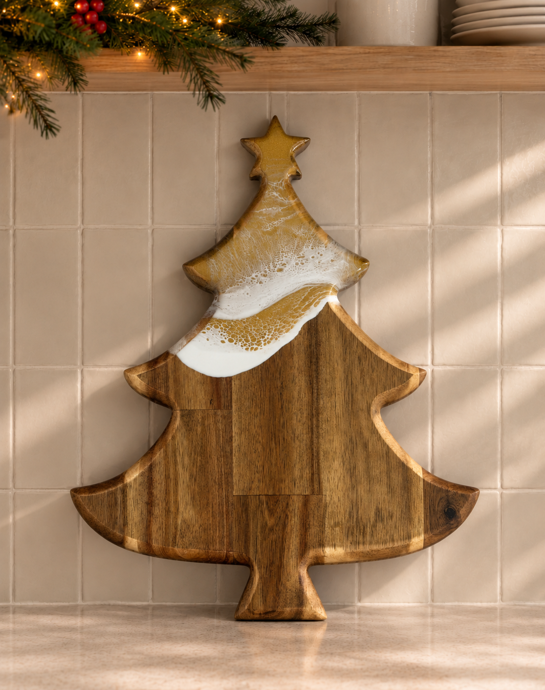 A Christmas tree-shaped wooden serving board with a gold glitter pattern on the top half and a white textured accent, displayed against a beige tiled wall with holiday greenery and string lights above.