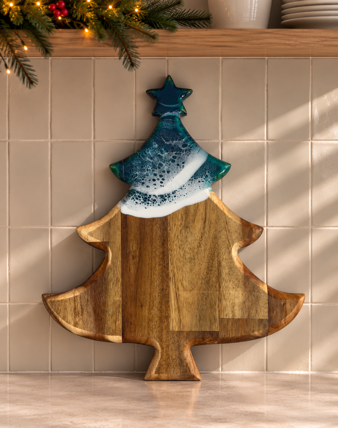 A Christmas tree-shaped wooden serving board with a glossy blue and white painted top section, displayed against a beige tiled wall with holiday greenery and string lights above.