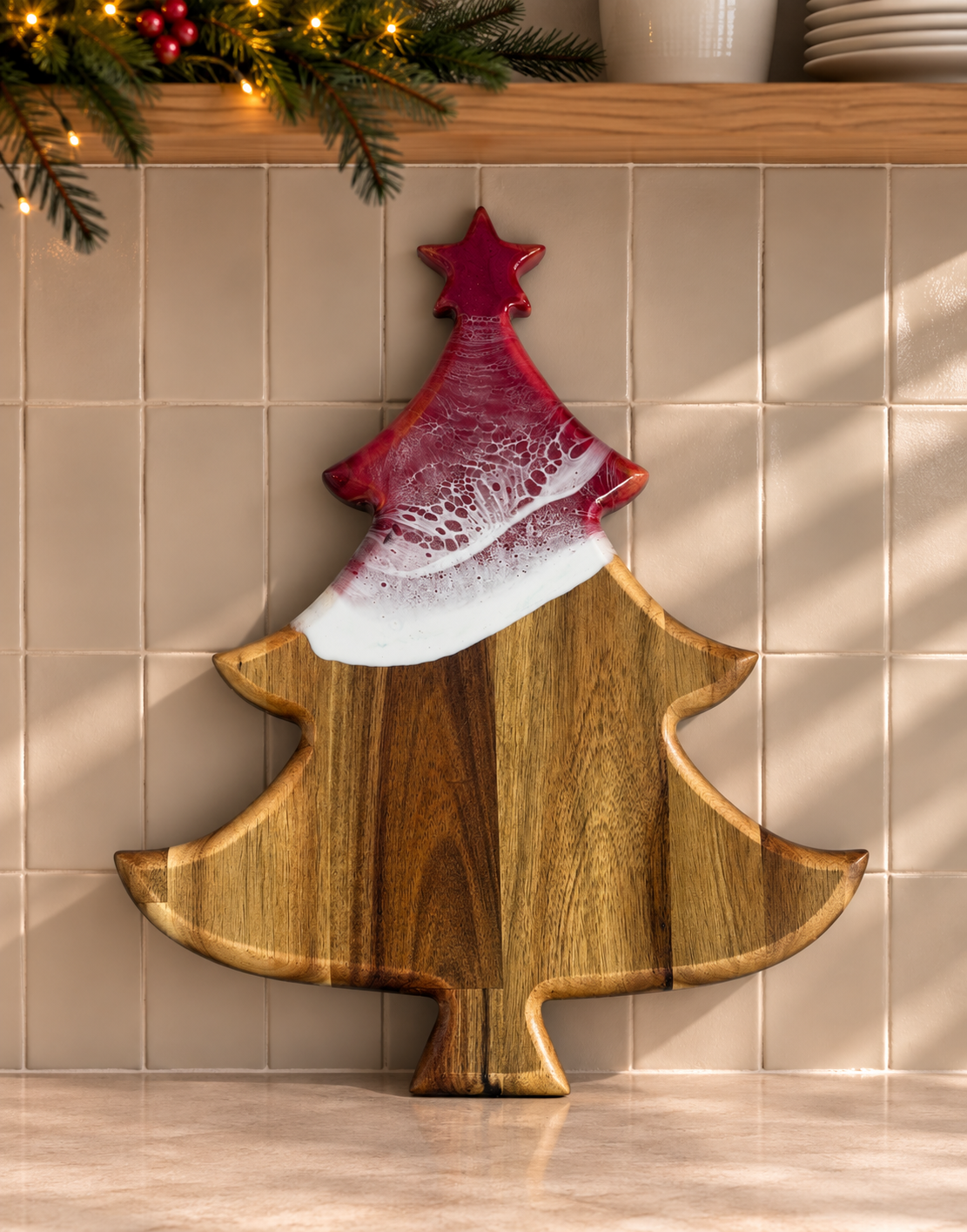 A Christmas tree-shaped wooden cutting board with a red and white marbled resin topper, displayed against a beige tiled wall with holiday greenery and warm lights in the background.