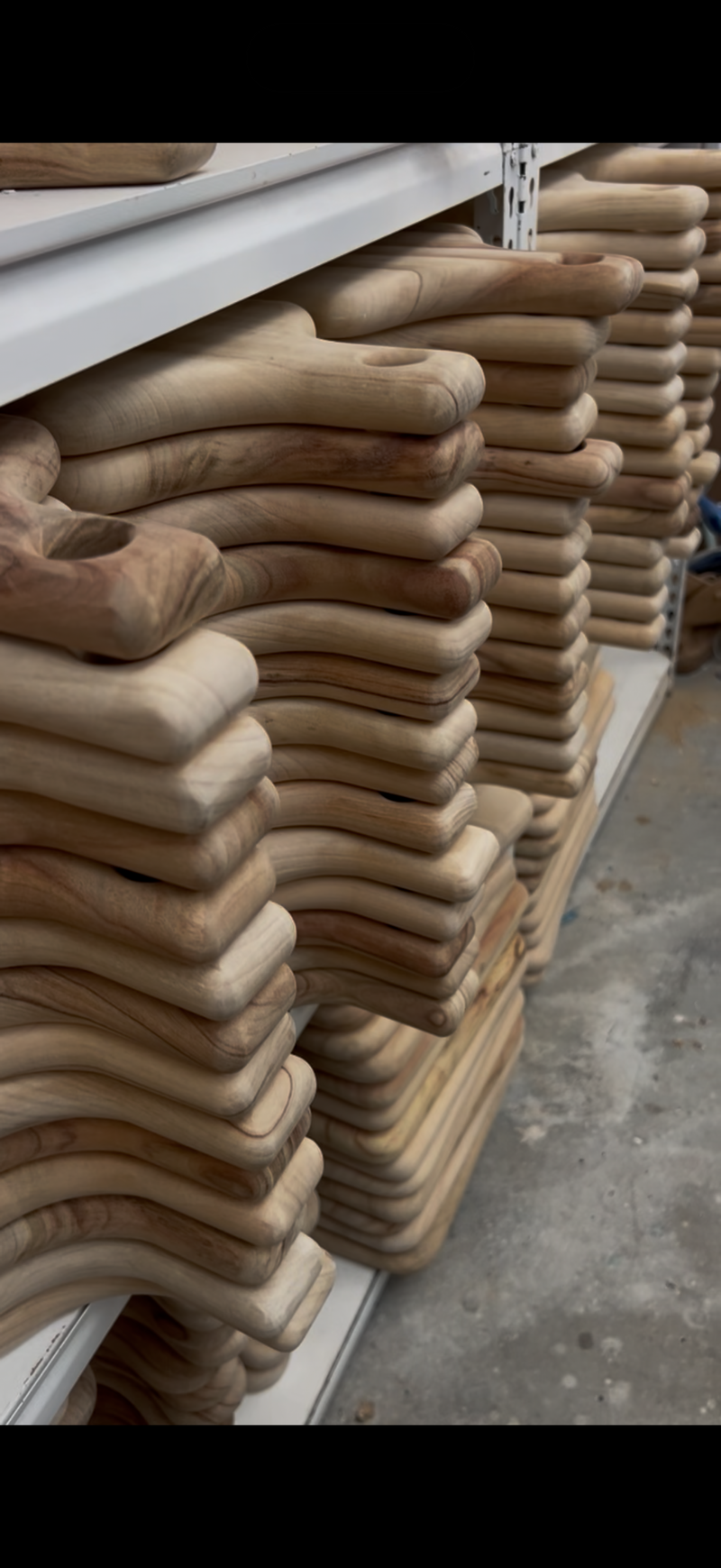 Stacks of pre cut wooden cutting boards with natural camphor laurel wood grain, stored on white shelves in what appears to be a warehouse or workshop.