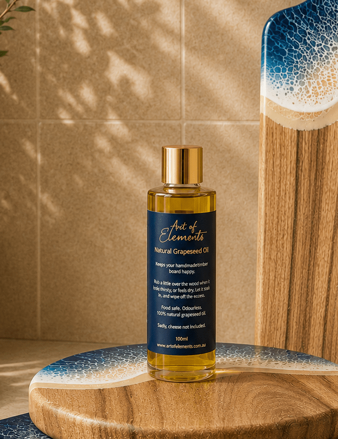 A bottle of Art of Elements Grapeseed Oil with a gold cap and dark navy label, styled on a wooden board against a beige tiled background with soft leaf shadows.