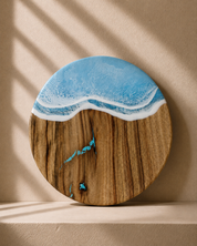 Round wooden serving board with a resin ocean wave design in light blue and white, displayed on a beige shelf with soft window shadow overlays.