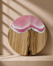 Round wooden serving board with a resin ocean wave design in pink and white, displayed on a beige shelf with soft window shadow overlays.