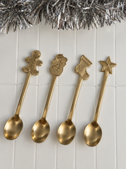 Four gold-toned decorative spoons with holiday-themed handles, including a gingerbread man, snowman, stocking, and star, arranged on a white paneled surface with silver tinsel at the top.