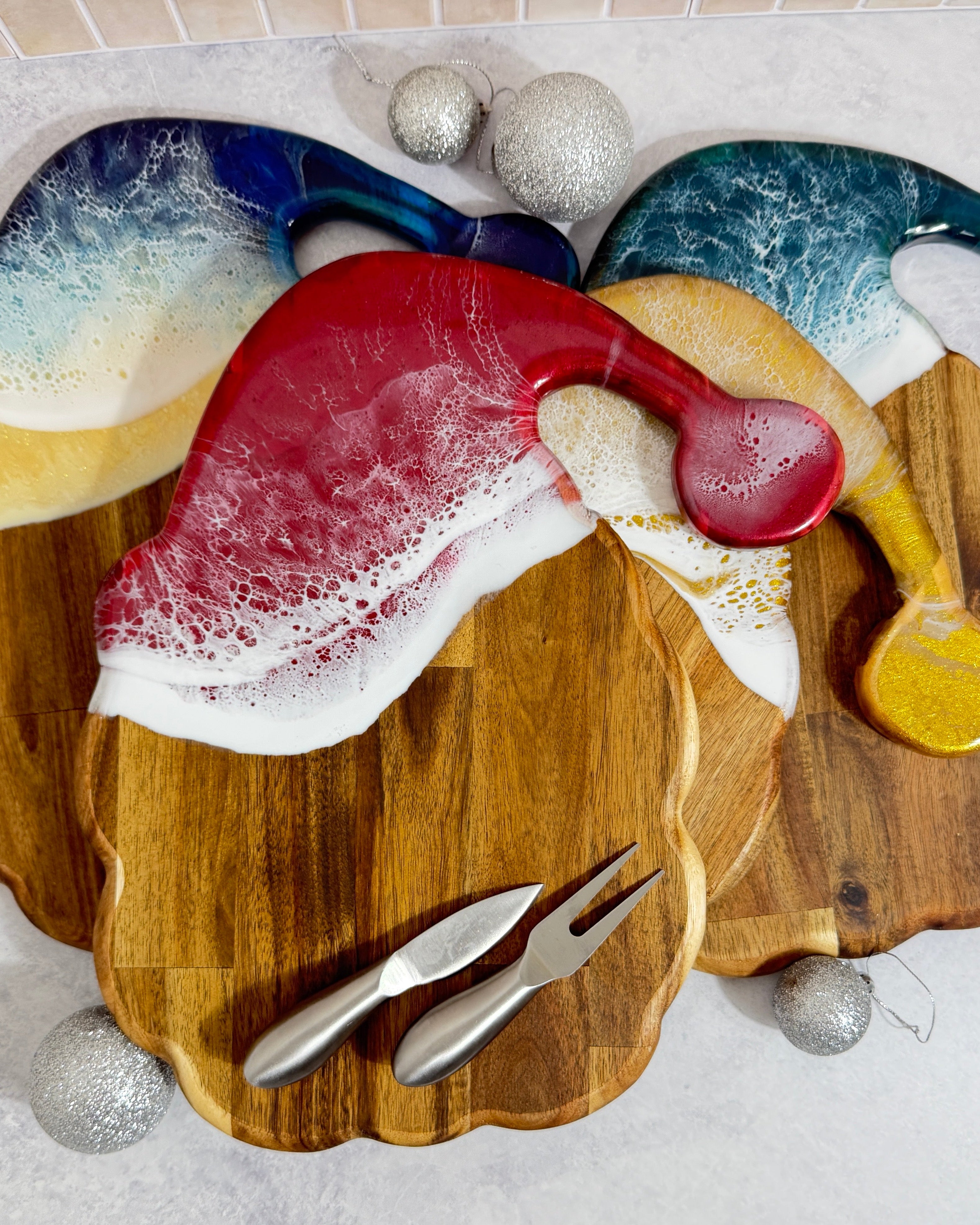 A festive holiday-themed serving board set on a light gray marble surface, featuring a scalloped wooden board with three colorful, Santa hat-shaped serving pieces in red, green, beach, and gold, accompanied by two silver metal utensils and silver holiday ornaments.