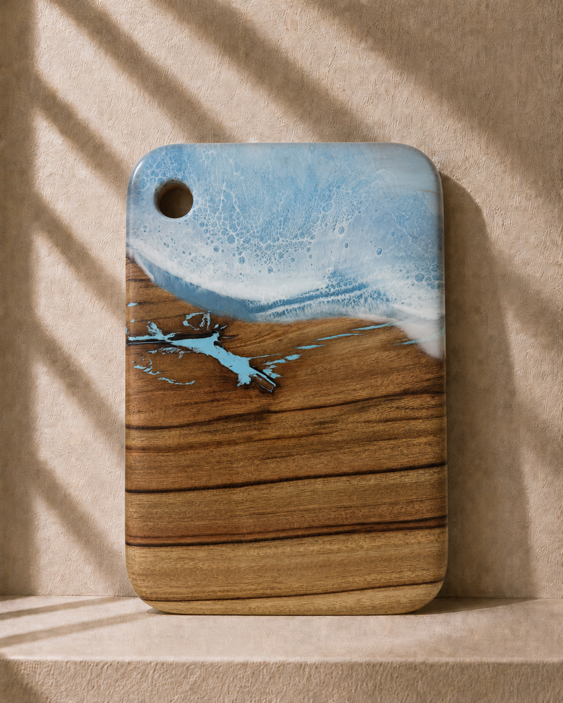 A rectangular camphor laurel wooden cutting board with rounded corners, featuring hand poured rolling resin wave design in light blue and white resin at the top and a natural light brown wood grain below. It is photographed standing upright on a matching beige surface against a textured beige wall with soft window shadow patterns.