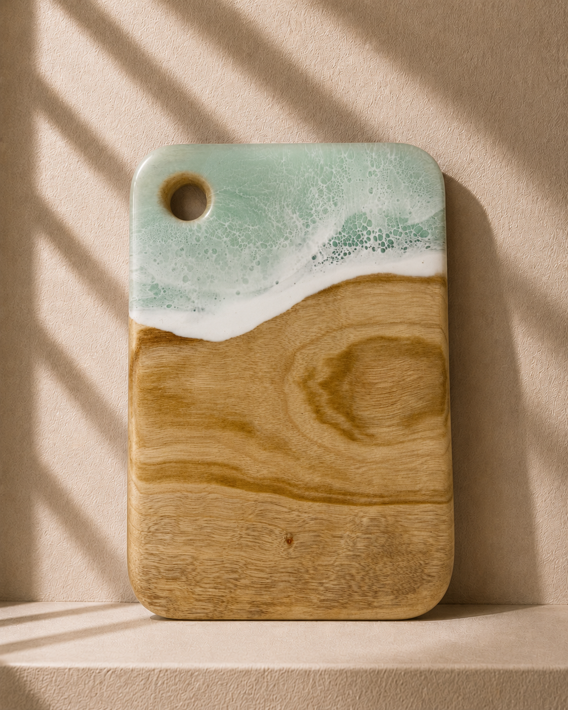 A rectangular camphor laurel wooden cutting board with rounded corners, featuring hand poured rolling resin wave design in mint green and white resin at the top and a natural light brown wood grain below. It is photographed standing upright on a matching beige surface against a textured beige wall with soft window shadow patterns.