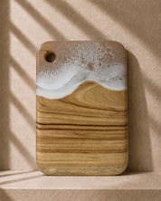 A rectangular camphor laurel wooden cutting board with rounded corners, featuring hand poured rolling resin wave design in beige and white resin at the top and a natural light brown wood grain below. It is photographed standing upright on a matching beige surface against a textured beige wall with soft window shadow patterns.