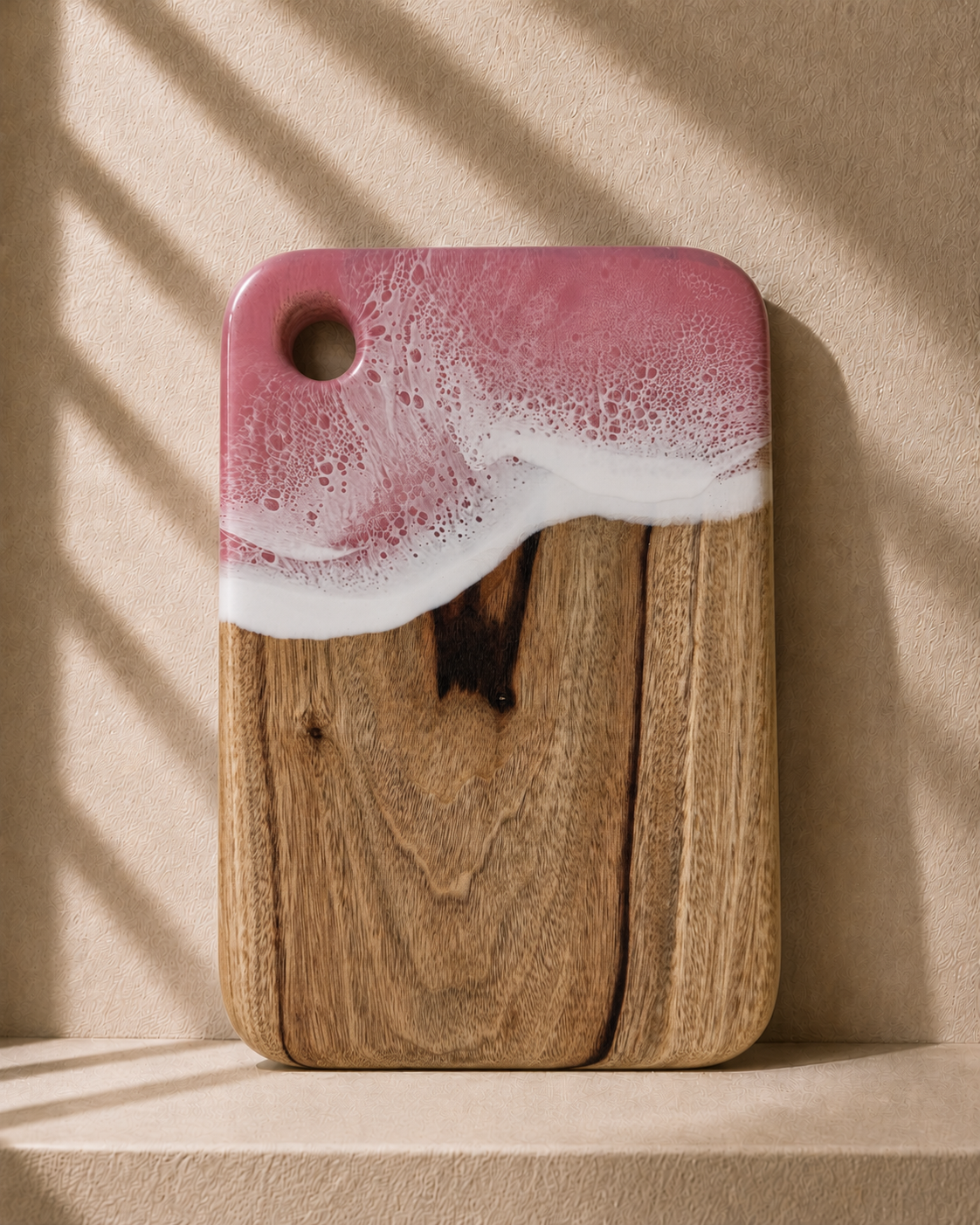 A rectangular camphor laurel wooden chopping board with rounded corners, featuring hand poured rolling resin wave design in pink and white resin at the top and a natural light brown wood grain below. It is photographed standing upright on a matching beige surface against a textured beige wall with soft window shadow patterns.