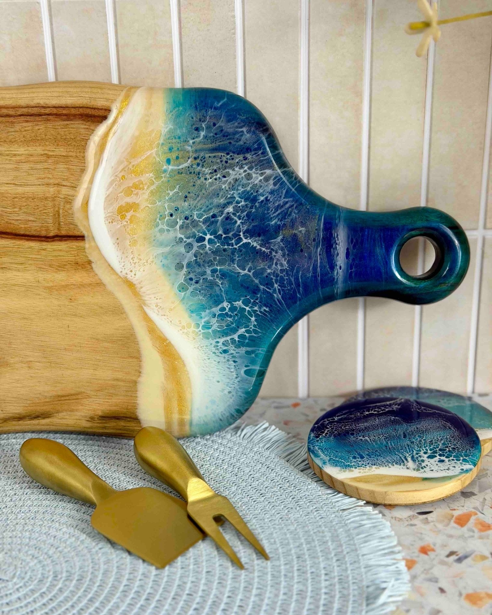 A decorative resin wooden serving board with a blue and gold resin waves, shown with matching gold-toned serving utensils and a small round coaster on a patterned surface.
