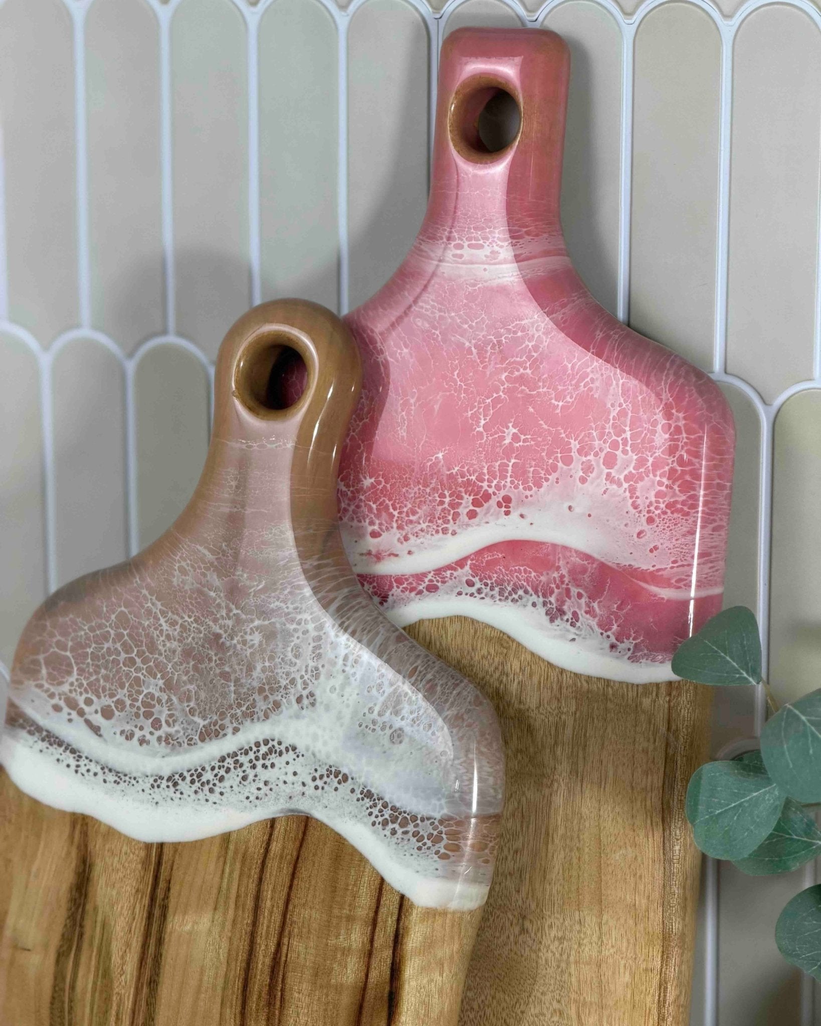 Two resin serving boards with a crackle glaze finish, one in pink and one in brown, displayed on a wooden cutting board against a tiled background.