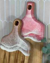 Two resin serving boards with a crackle glaze finish, one in pink and one in brown, displayed on a wooden cutting board against a tiled background.
