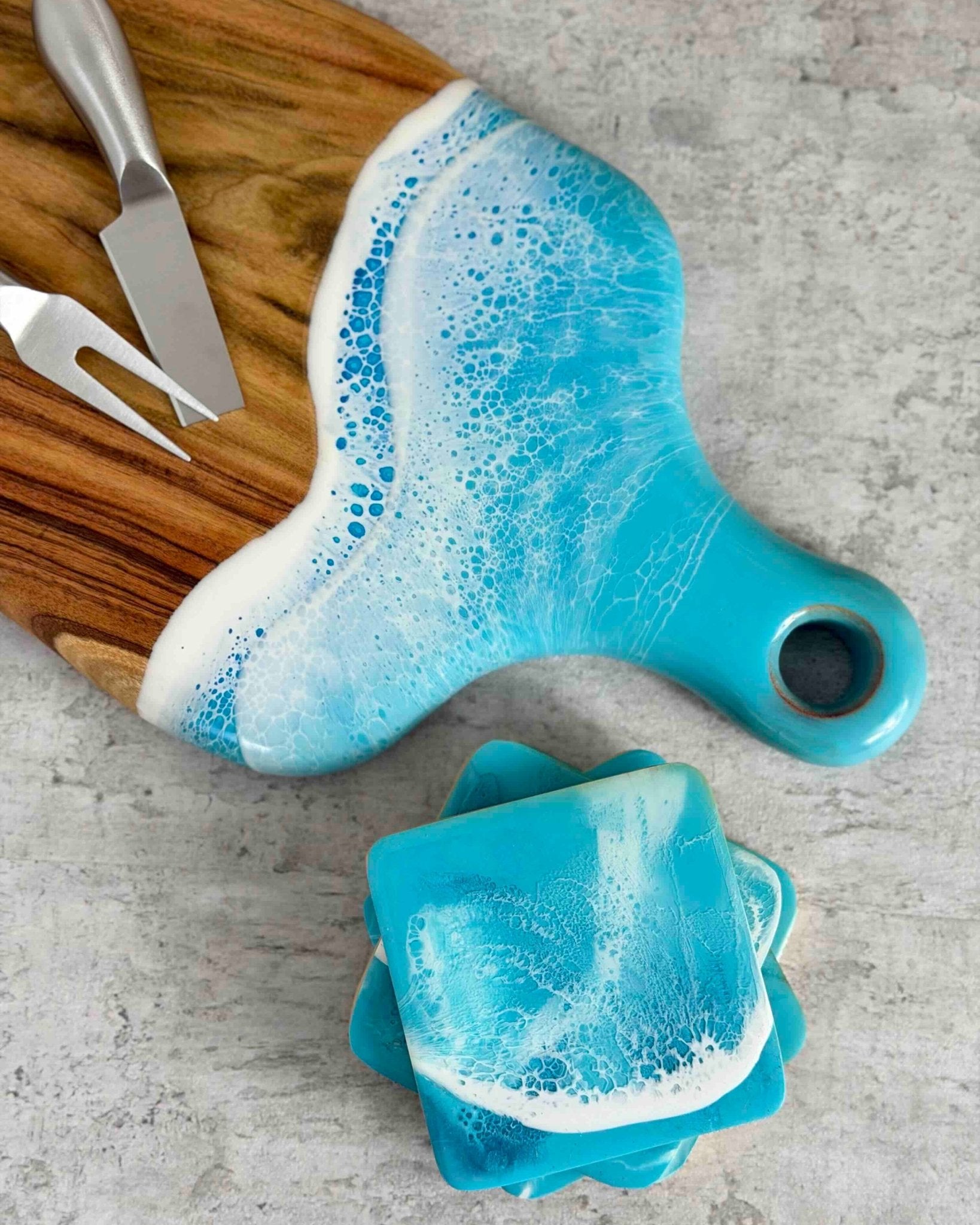 A cheeseboard with light pale blue rolling ocean resin waves covering the handle. Matching coasters lay by its side and silver cheeseknives sit on the board.