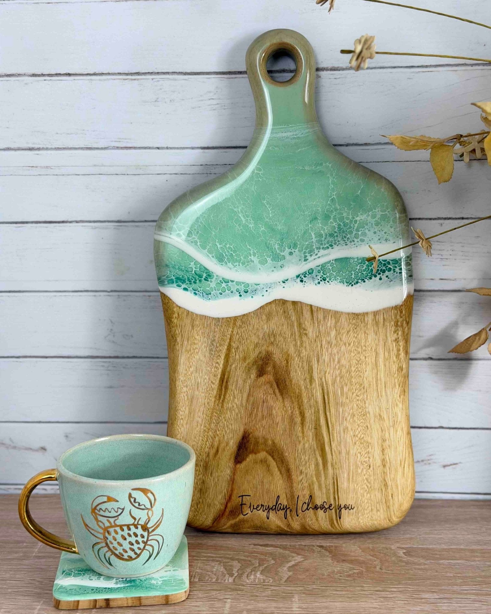 A wooden serving board with a mint green and white wave on the top half covering the handle, styled on a wooden surface next to a matching mint green mug with a gold crab illustration.