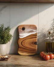 A wavy-edged wooden cutting board with a resin wave pattern in warm brown and tan tones, leaning against a light marble kitchen backsplash. The board features a circular hanging hole and is styled on a wooden countertop with fresh tomatoes, a potted herb, and an oil bottle.