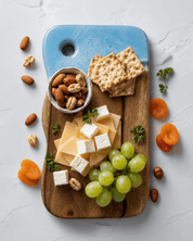 A styled flat-lay of a cutting board with a charcuterie board setup, featuring crackers, cheese cubes, green grapes, nuts, and dried apricots on a light gray marble surface.