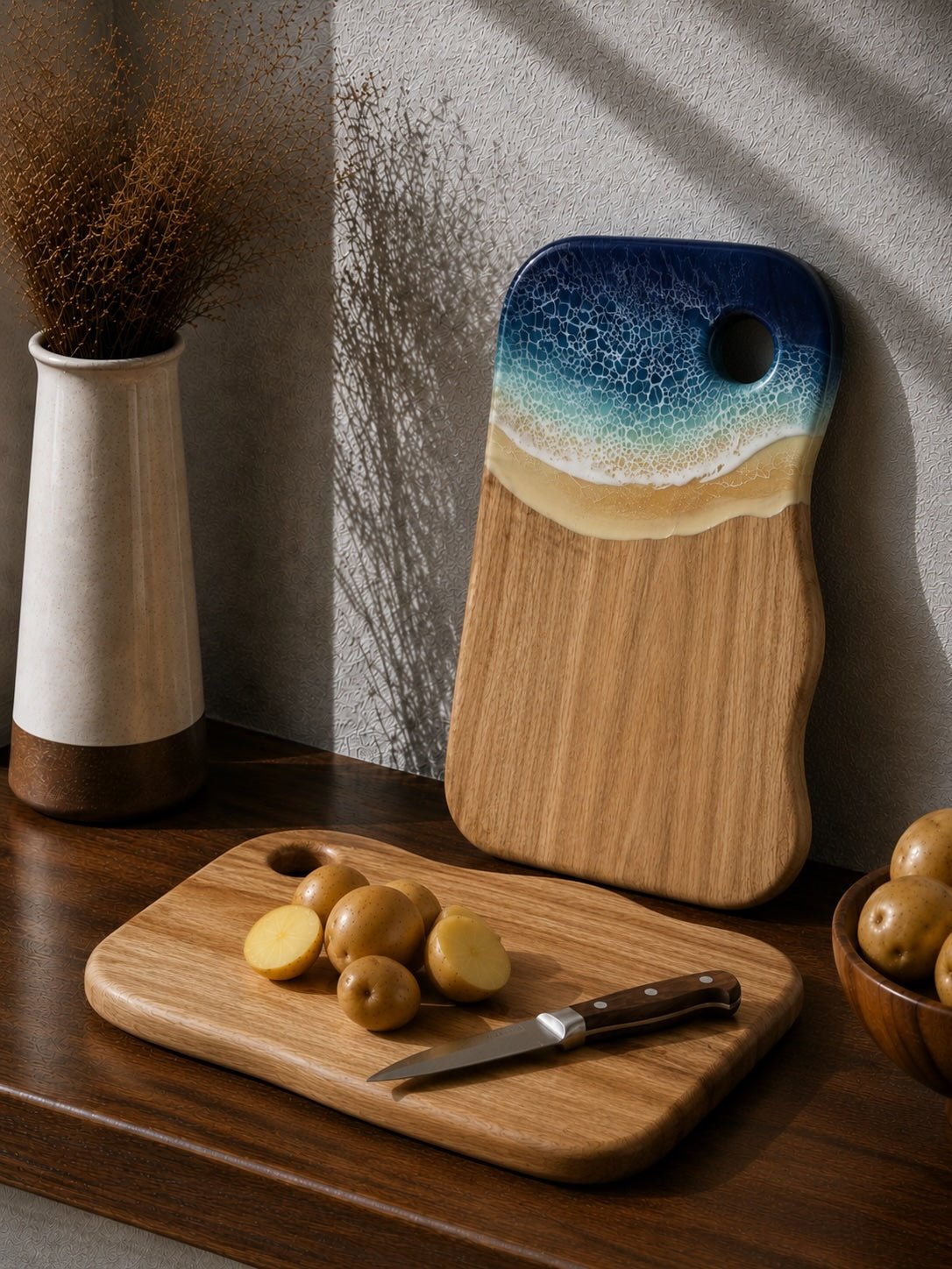 Two wooden cutting boards with a beach-themed design, one standing upright leaning against a wall and the other lying flat on a wooden table with potatoes and a knife. The boards feature a natural wood grain body with a painted wave design in blue, teal, and beige at the top.