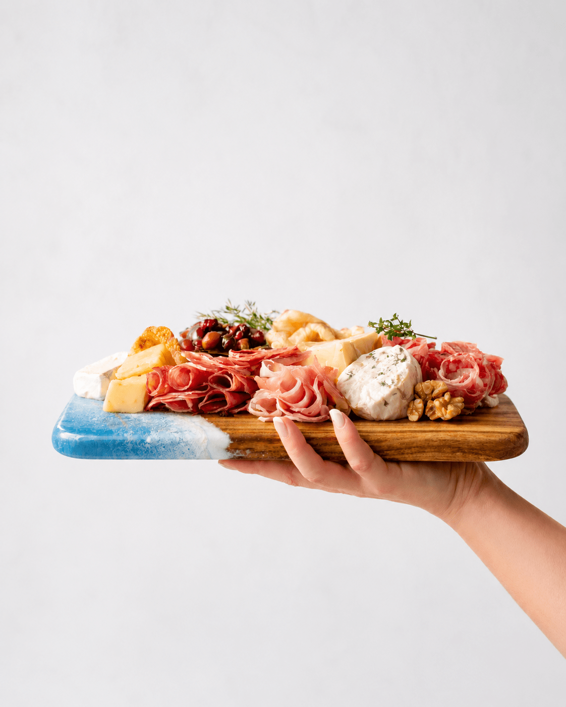 Hand holding the mini charcuterie board with various meats, cheeses, and fruits on a light gray background