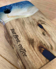 A close-up of a camphor laurel cutting board with a blue, sand coloured resin waves and the engraved text 'But first, coffee' on a light wood surface.