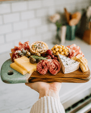 A charcuterie board with cheese, cured meats, crackers, olives, and nuts held by a hand in a bright kitchen setting.