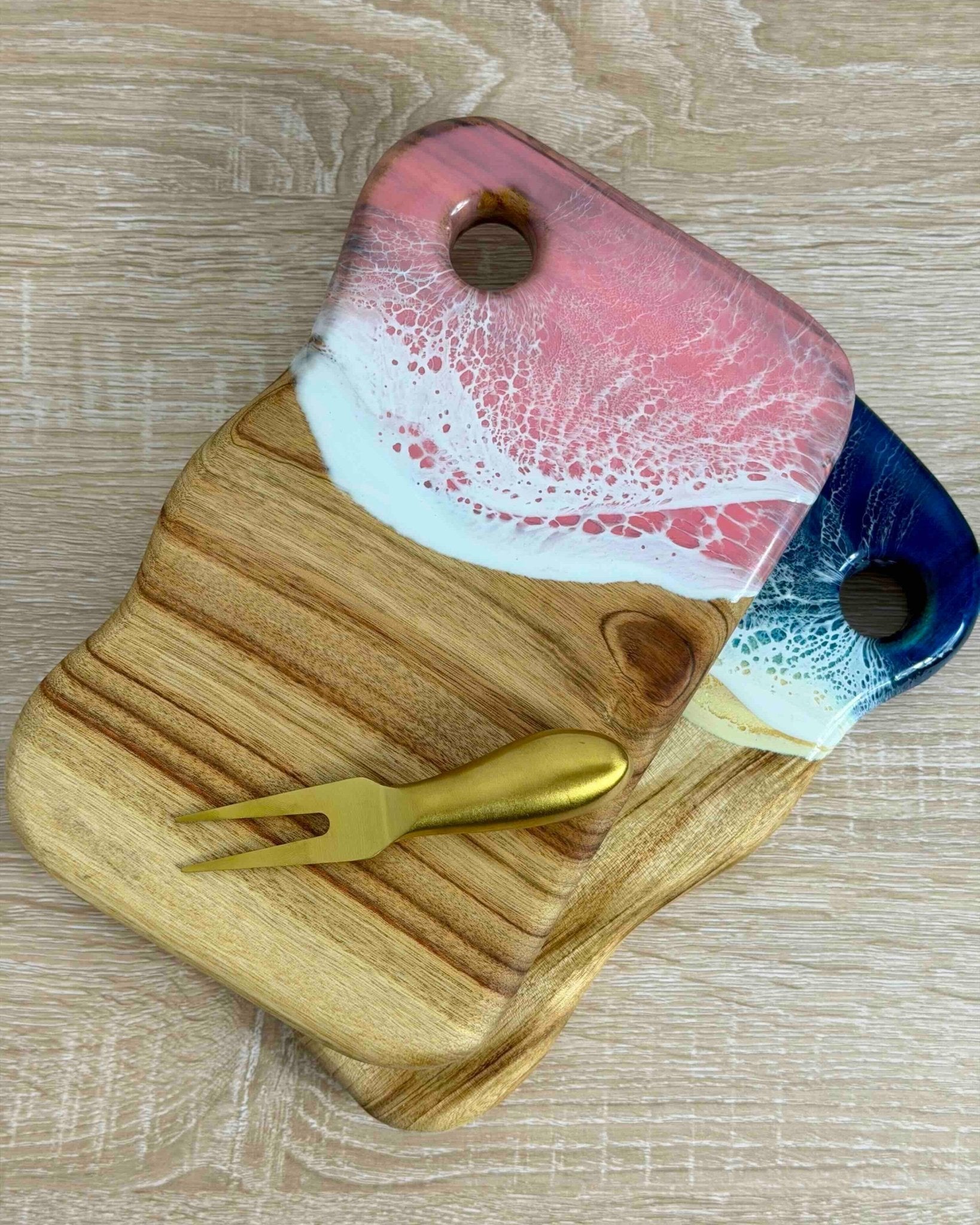 Mini cutting board with a pink and beach resin waves and a gold cheese knife on a wooden surface.