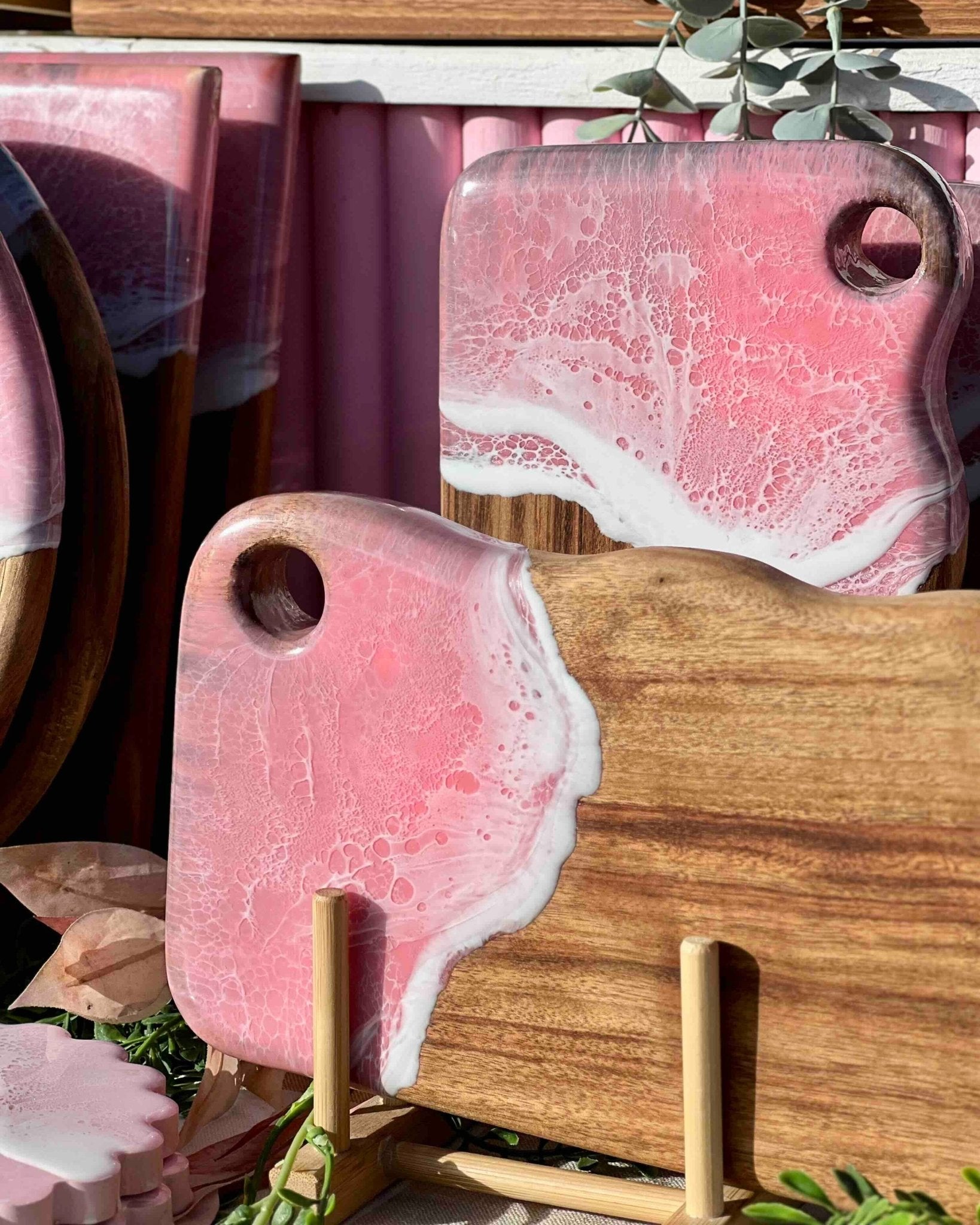 Pink and white epoxy resin cutting boards with hanging holes displayed in a stand in natural sunlight at a market