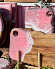 Pink and white epoxy resin cutting boards with hanging holes displayed in a stand in natural sunlight at a market