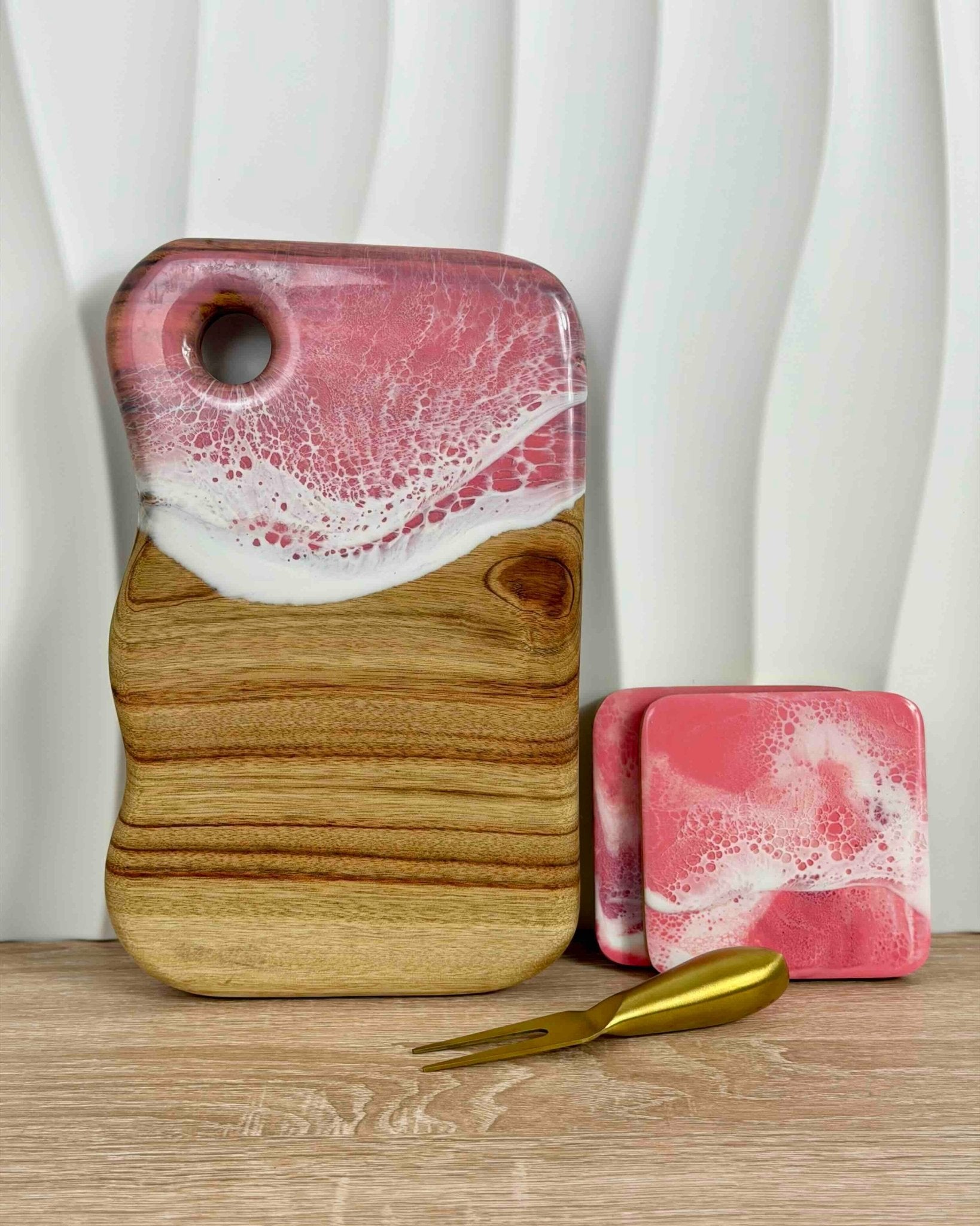 A pink and white epoxy resin wooden cutting board with a cutout in the top left, shown on a wooden surface with two matching pink and white resin coasters and a gold cheese knife against a white wavy background.