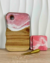 A pink and white epoxy resin wooden cutting board with a cutout in the top left, shown on a wooden surface with two matching pink and white resin coasters and a gold cheese knife against a white wavy background.