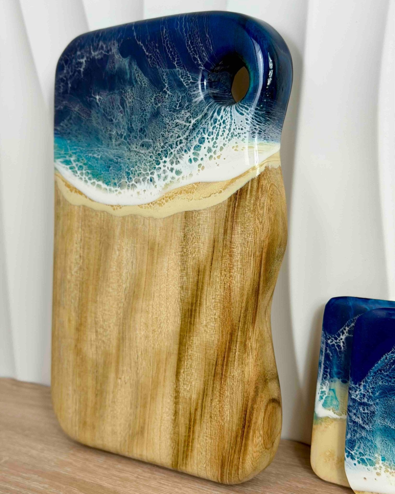 Wooden cutting board with a wavy edge, and a hanging hole cutout, with blue and white rolling resin waves