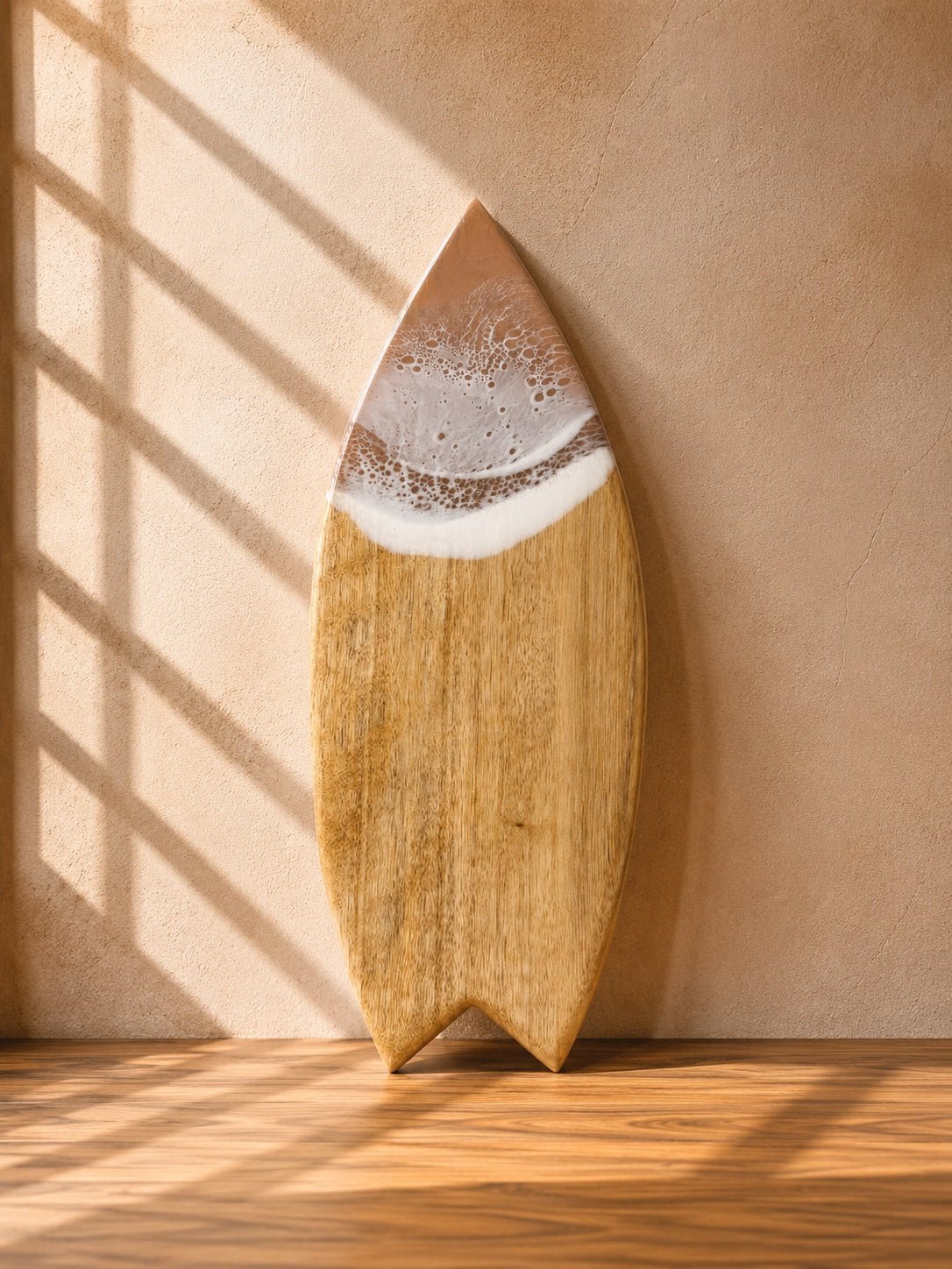 A surfboard-shaped wooden cutting board with a camphor laurel wood grain and a beige resin wave, leaning against a textured wall with diagonal window shadow patterns.