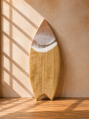 A surfboard-shaped wooden cutting board with a camphor laurel wood grain and a beige resin wave, leaning against a textured wall with diagonal window shadow patterns.
