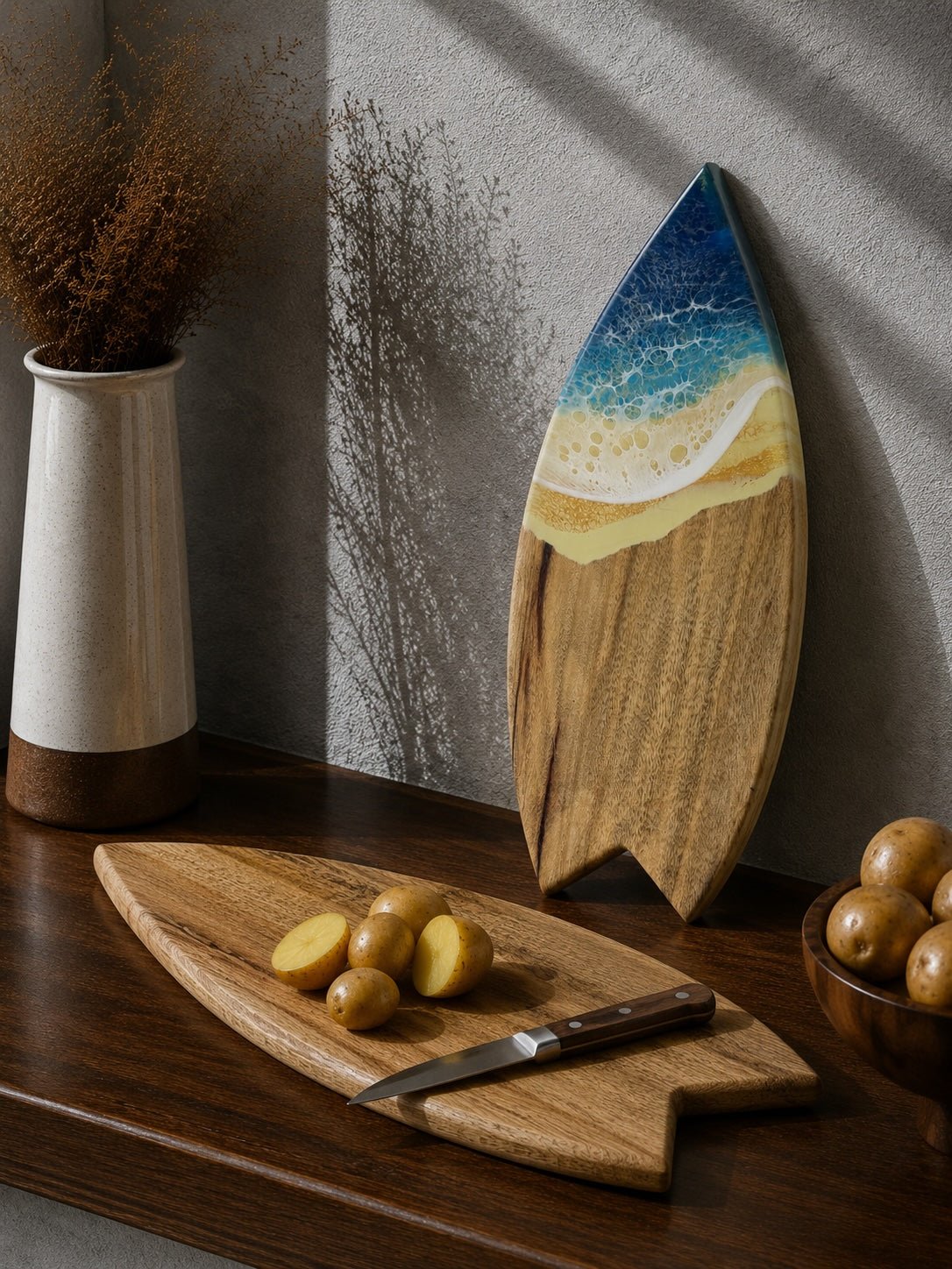 A surfboard-shaped wooden cutting board with a blue ocean and sandy beach design, displayed on a dark wooden table next to a matching wooden potato board with a knife and potatoes.