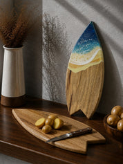 A surfboard-shaped wooden cutting board with a blue ocean and sandy beach design, displayed on a dark wooden table next to a matching wooden potato board with a knife and potatoes.