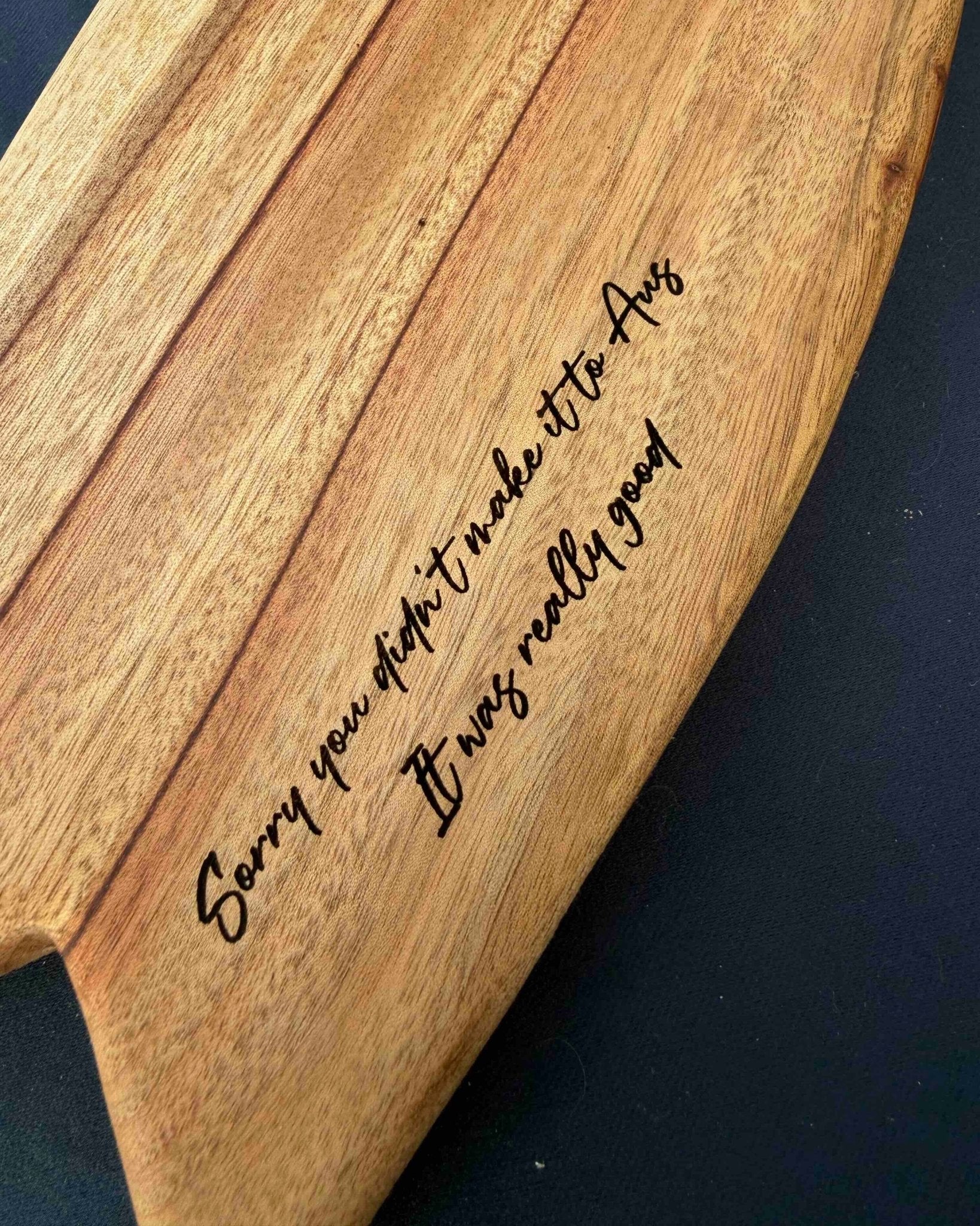 A close-up of a curved mini surfboard shaped cheese board, featuring engraved cursive text that reads 'Sorry you didn't make it to Aes It was really good'. The board is shown on a dark, solid background.