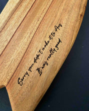A close-up of a curved mini surfboard shaped cheese board, featuring engraved cursive text that reads 'Sorry you didn't make it to Aes It was really good'. The board is shown on a dark, solid background.