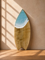 Wooden surfboard-shaped chopping board light blue resin waves on the top leaning on a kitchen wall in natural lighting